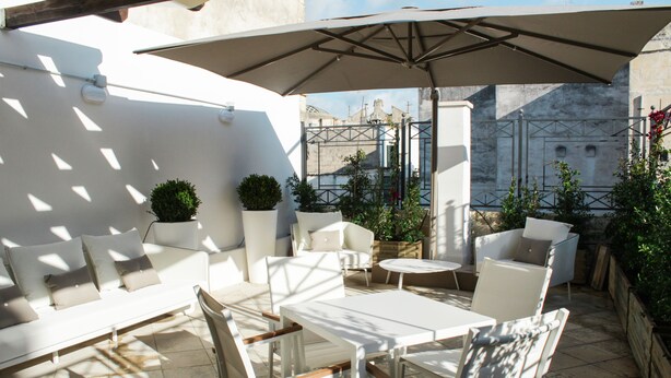 Un patio exterior con mobiliario blanco, incluyendo un sofá y sillas, bajo un paraguas grande.
