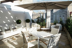 Un patio exterior con mobiliario blanco, incluyendo un sofá y sillas, bajo un paraguas grande.