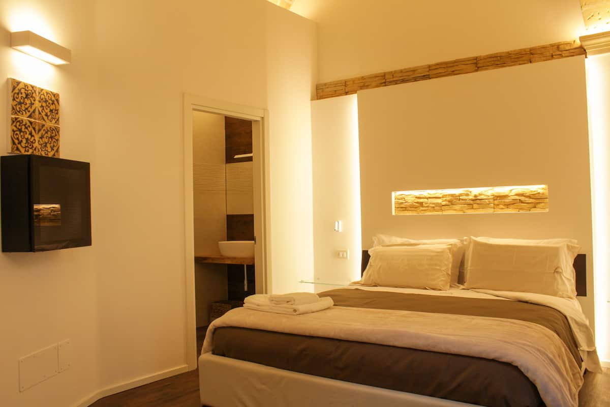 A bedroom with a bed, a nightstand, a wall-mounted TV, and a bathroom visible through an open door.