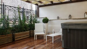 Un patio exterior con suelo de madera, sillas blancas, un jacuzzi y plantas en macetas.