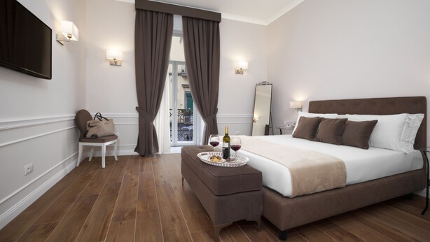 A modern hotel room with a large bed, a bedside table with a tray of snacks and wine, a flat-screen TV, and a view of the outside balcony. - La Fontanina Suites (Naples)