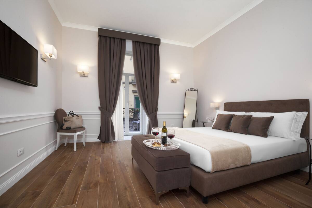 A modern hotel room with a large bed, a bedside table with a tray of snacks and wine, a flat-screen TV, and a view of the outside balcony.