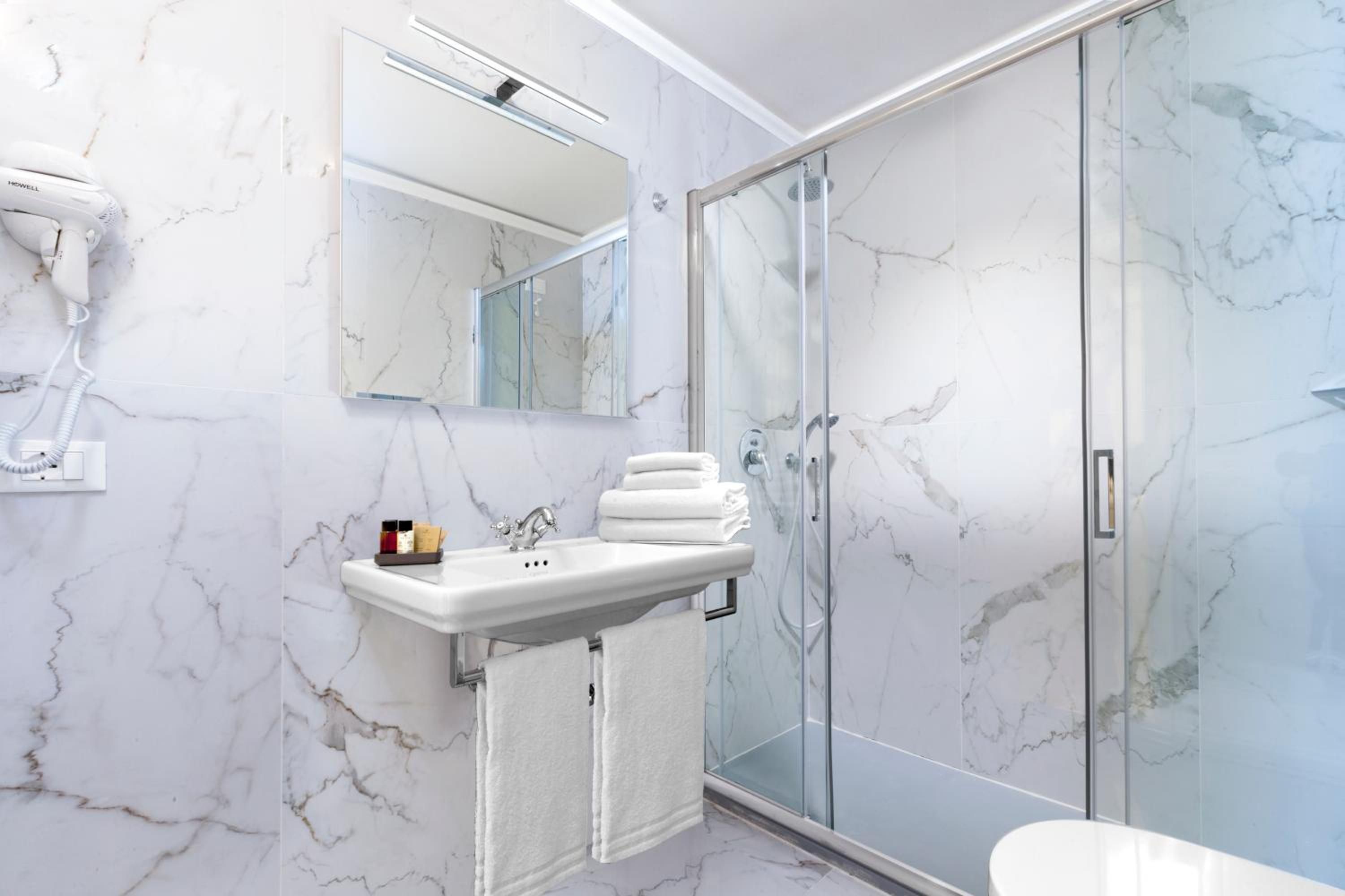 A bathroom with a marble wall, a sink, a mirror, a hairdryer, and a glass-enclosed shower.