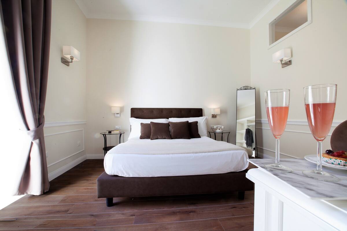 A neatly arranged bedroom with a large bed, bedside tables, a mirror, and two glasses of pink drink on a marble table.