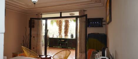 A room with a bed, two chairs, a TV, and a view of palm trees through an open door.