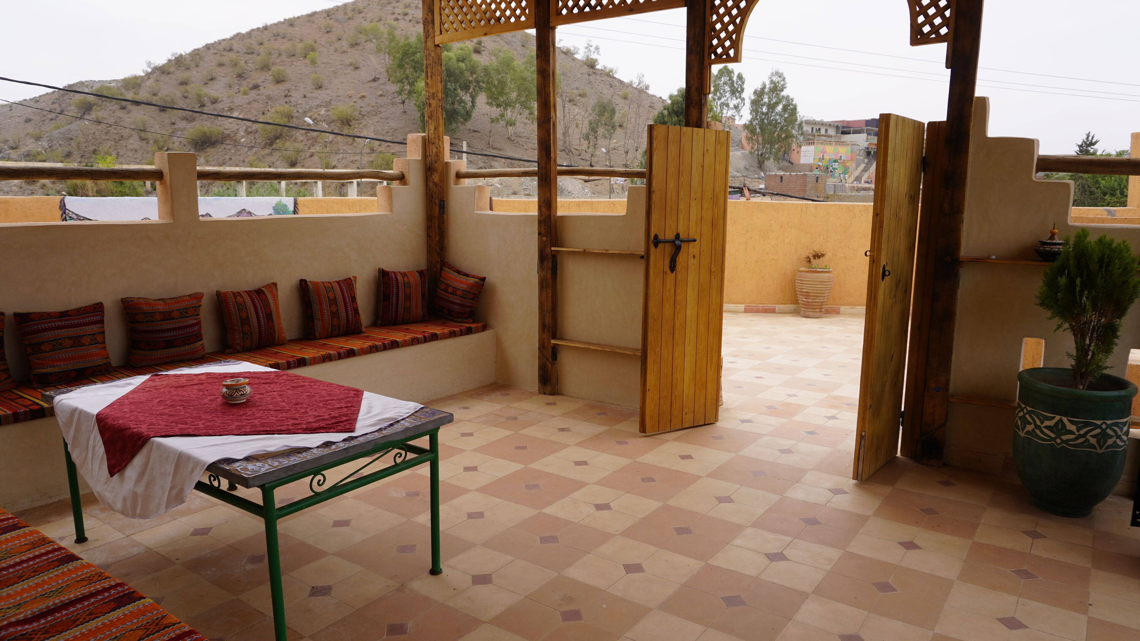 Una habitación con una cama, un banco con cojines, una puerta de madera y vistas a una colina.