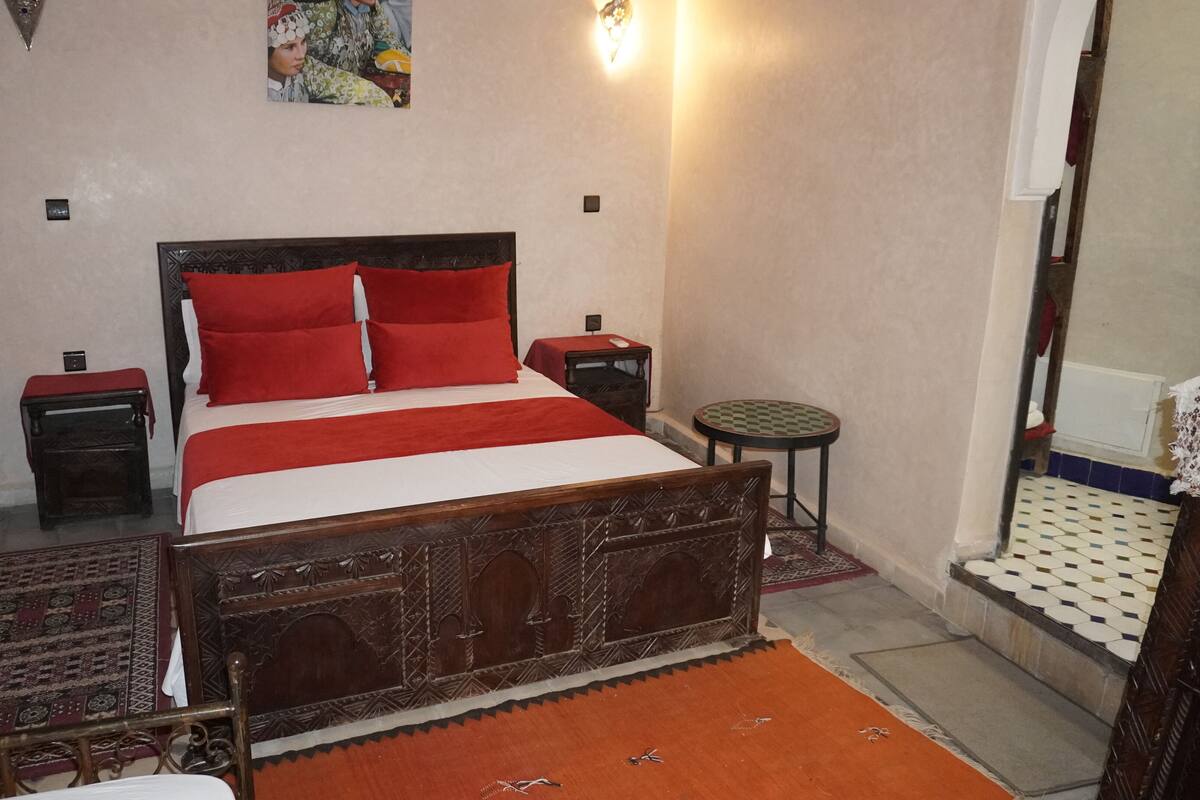 A bedroom with a wooden bed, red pillows, a small bedside table, a round table, and a patterned rug.
