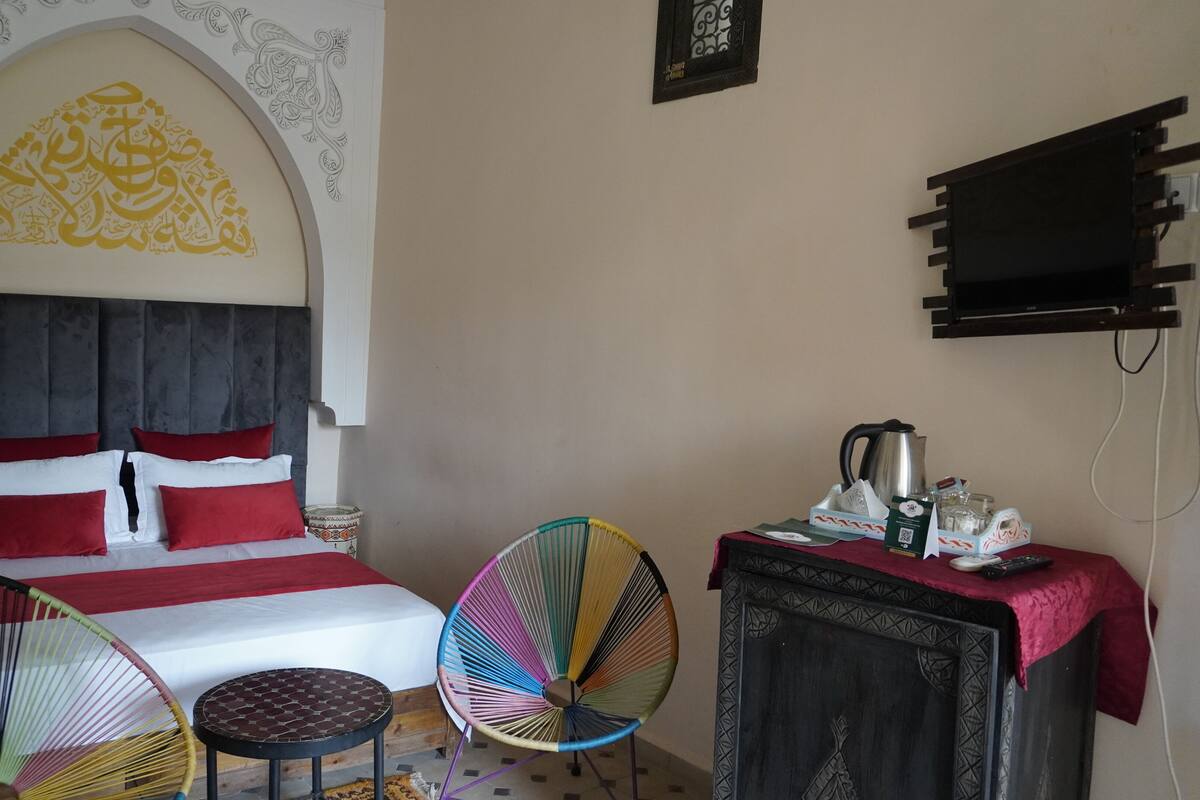 A room with a bed, two colorful chairs, a small table, a cabinet with a kettle, and a television.