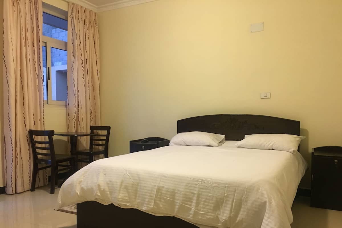 A bedroom with a large bed, two bedside tables, a small table with chairs, and a window with curtains.