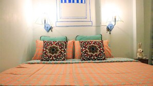 A bed with an orange patterned bedspread and two decorative pillows, a wall with a blue and white geometric design, and a small table with a teddy bear.