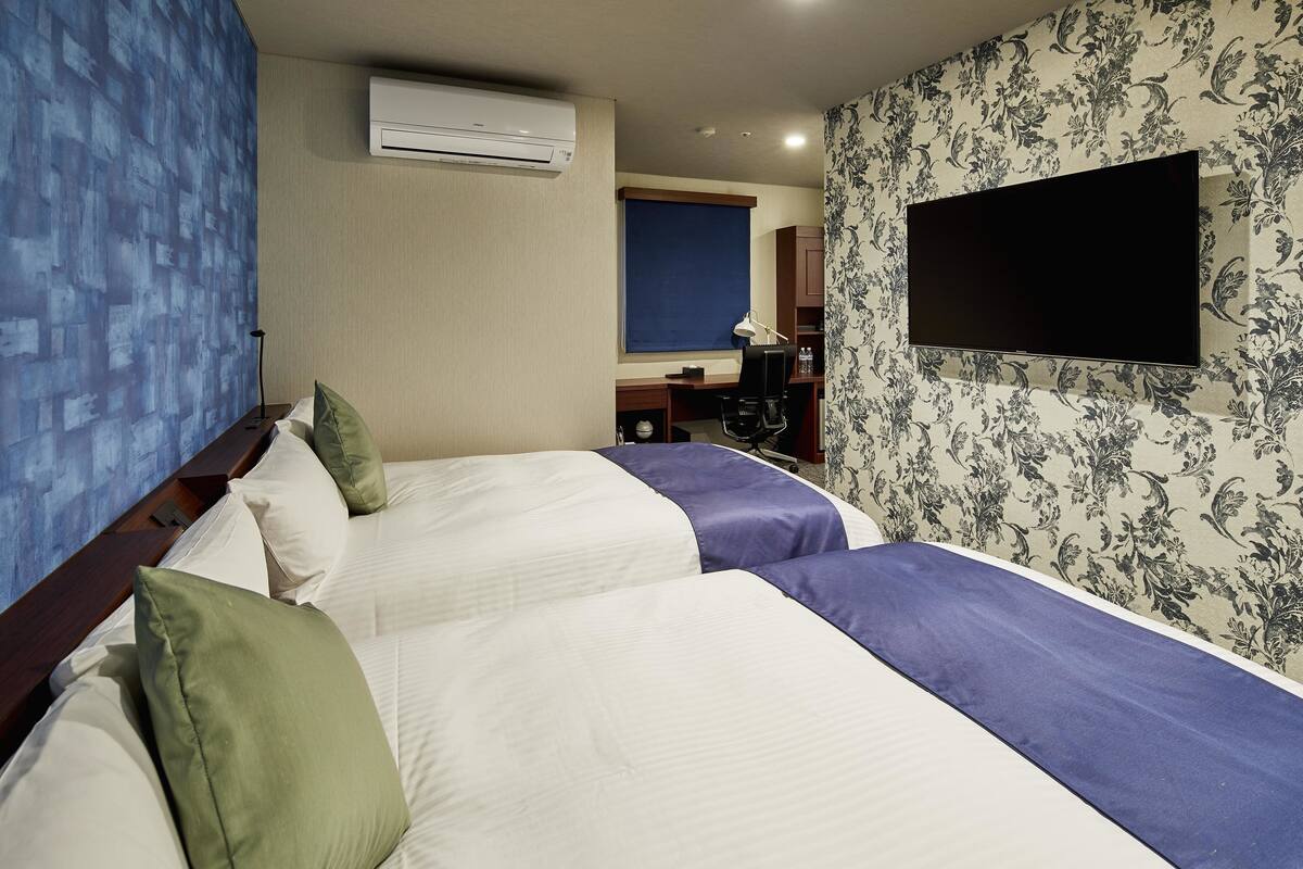 A hotel room with two beds, a TV mounted on the wall, an air conditioning unit, and a desk with a chair.