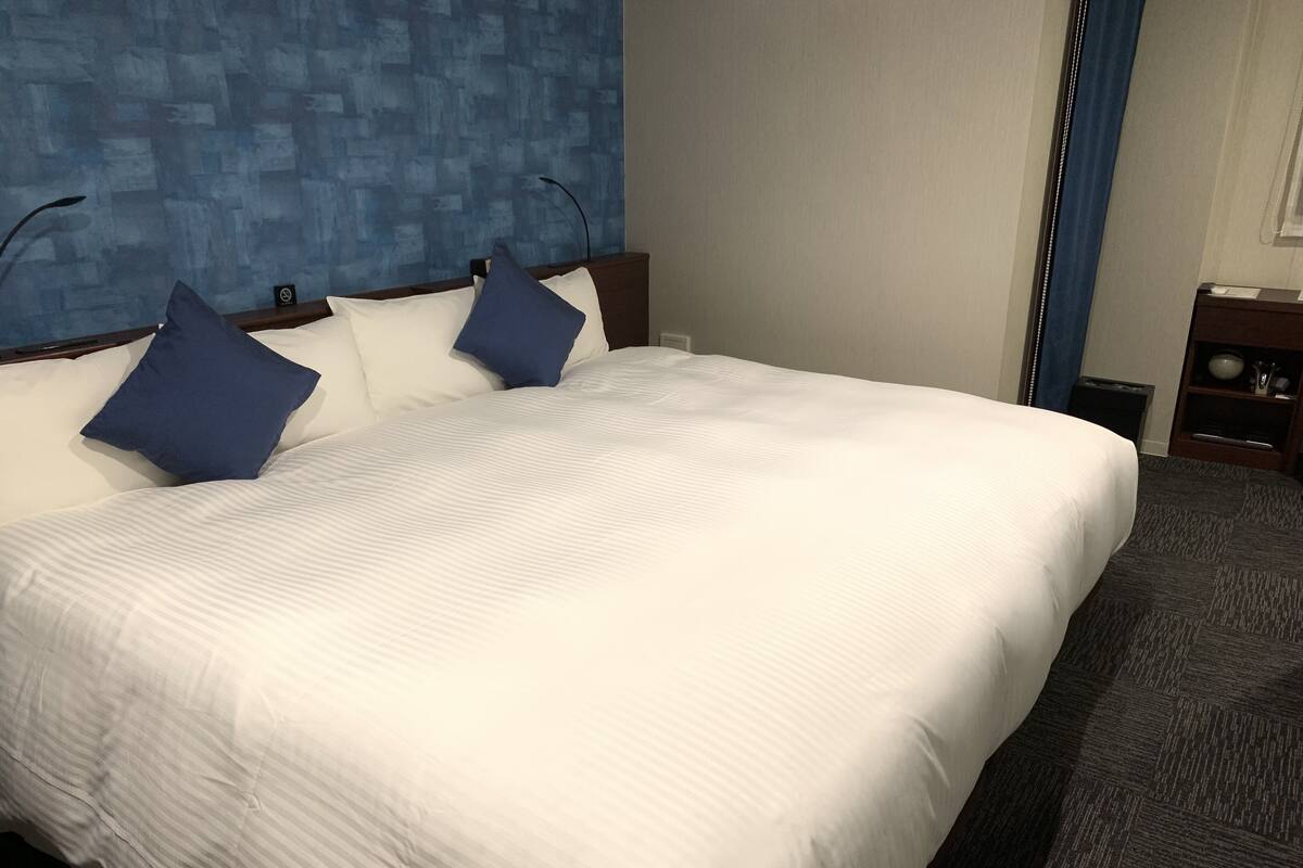 A hotel room with a large bed, a blue textured wall, and a small bedside table with a lamp.