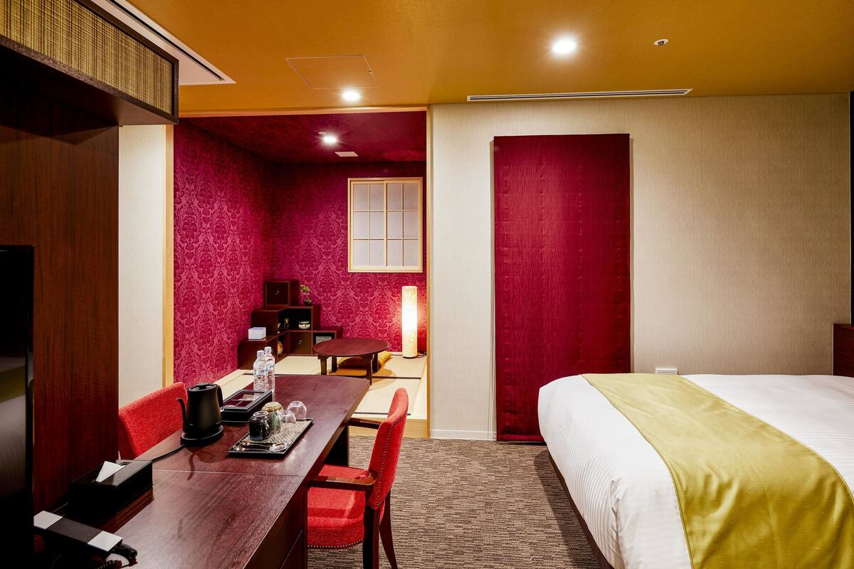 A hotel room with a bed, a desk with a chair, a table with a chair, a lamp, and a window.