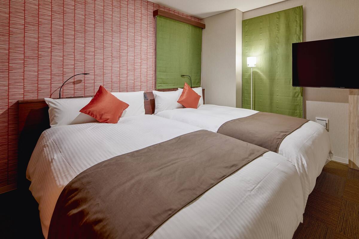 A hotel room with two beds, a flat-screen TV, a bedside lamp, and a window with a green curtain.