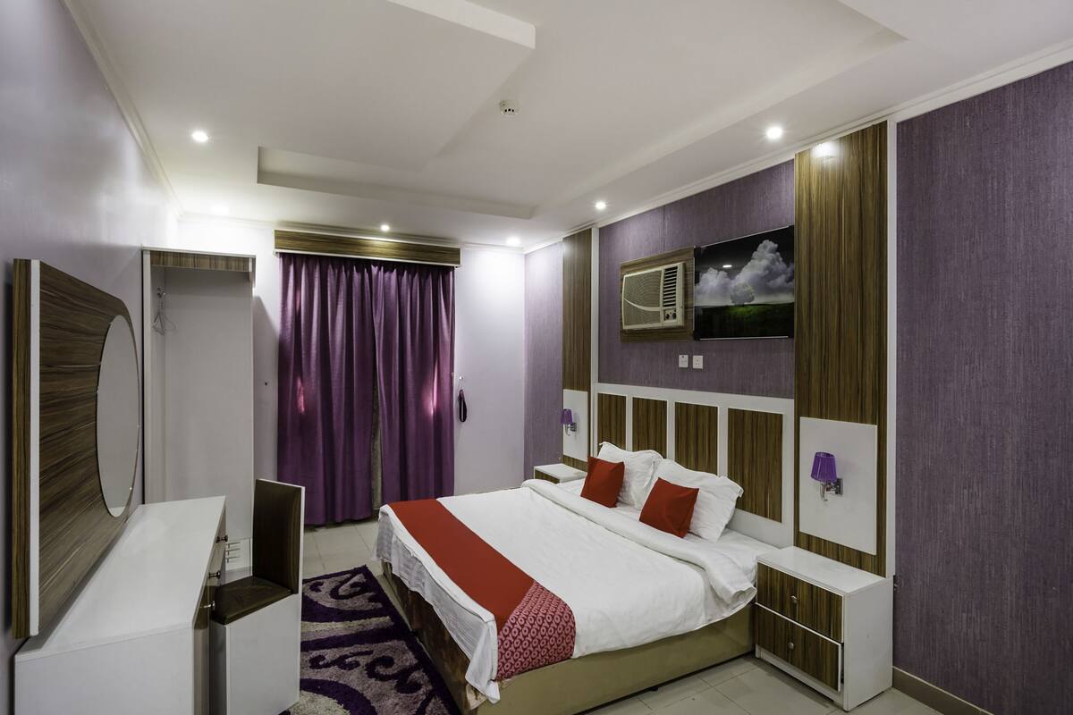 A modern hotel room with a large bed, a vanity with a mirror, a bedside table, and a wall-mounted air conditioning unit.