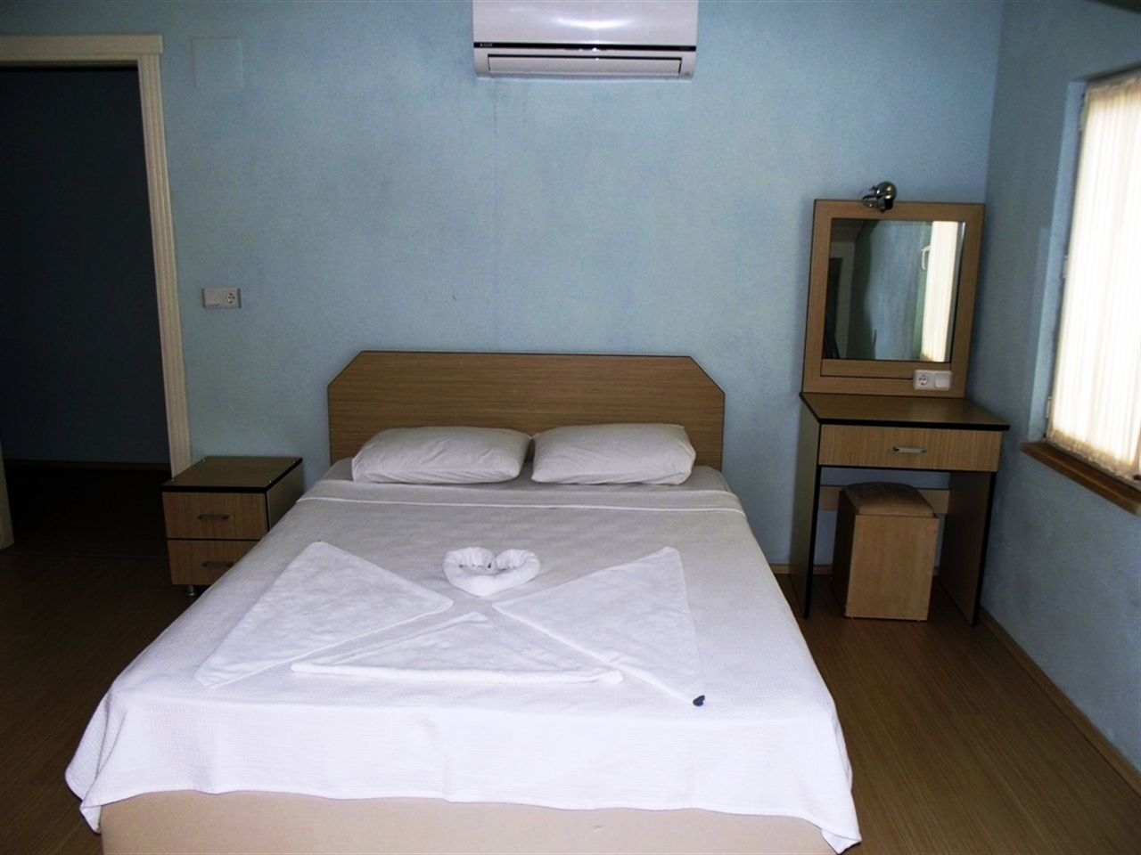 Una cama bien hecha con una toalla dispuesta en forma de corazón, una mesita de noche, un espejo y un equipo de aire acondicionado en la pared.