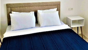 A hotel room with a bed, white pillows, a blue bedspread, a wooden headboard, a white nightstand, and a wall-mounted air conditioner. - Dalyan Gurkan Apart Hotel (Ortaca)