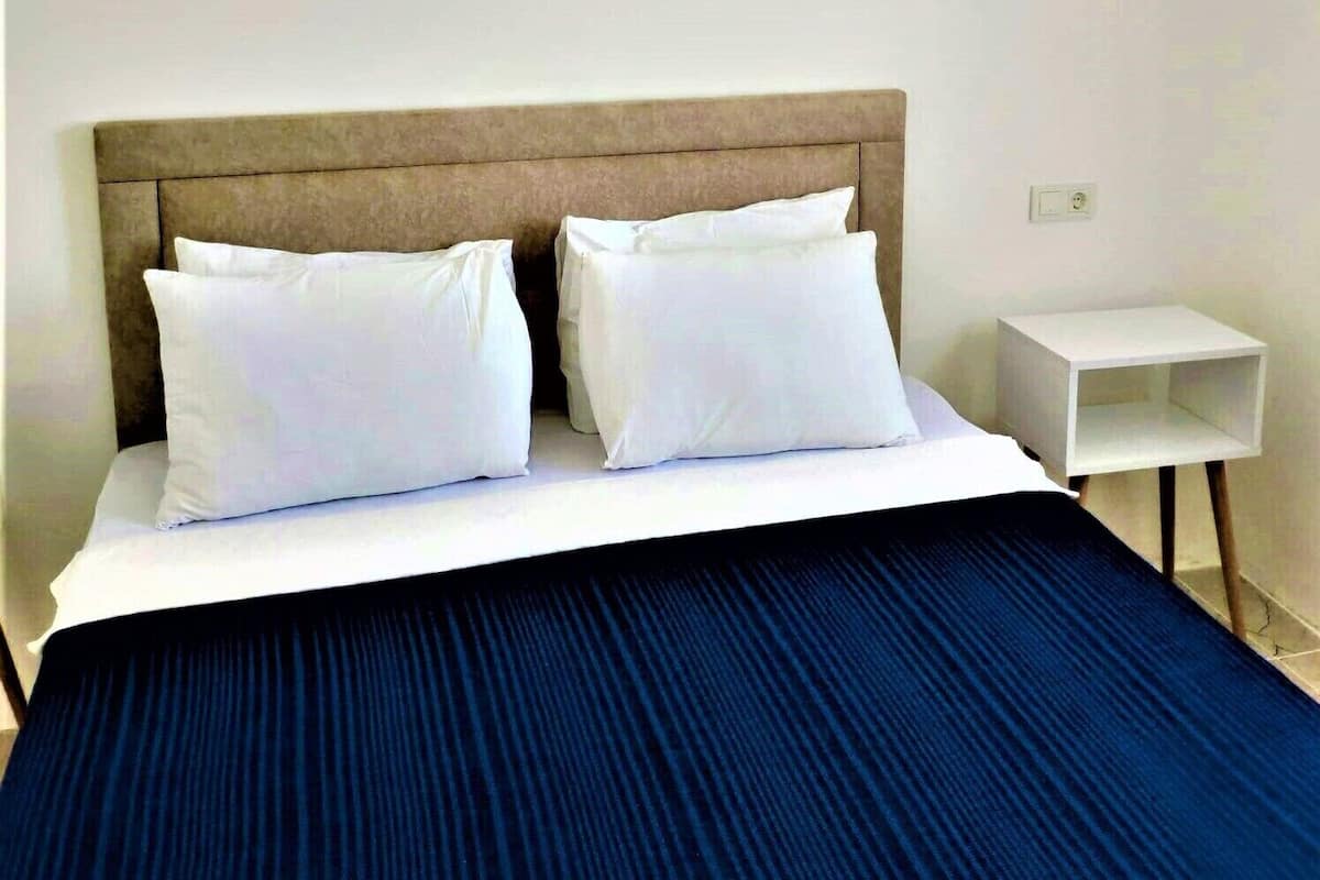 A hotel room with a bed, white pillows, a blue bedspread, a wooden headboard, a white nightstand, and a wall-mounted air conditioner.