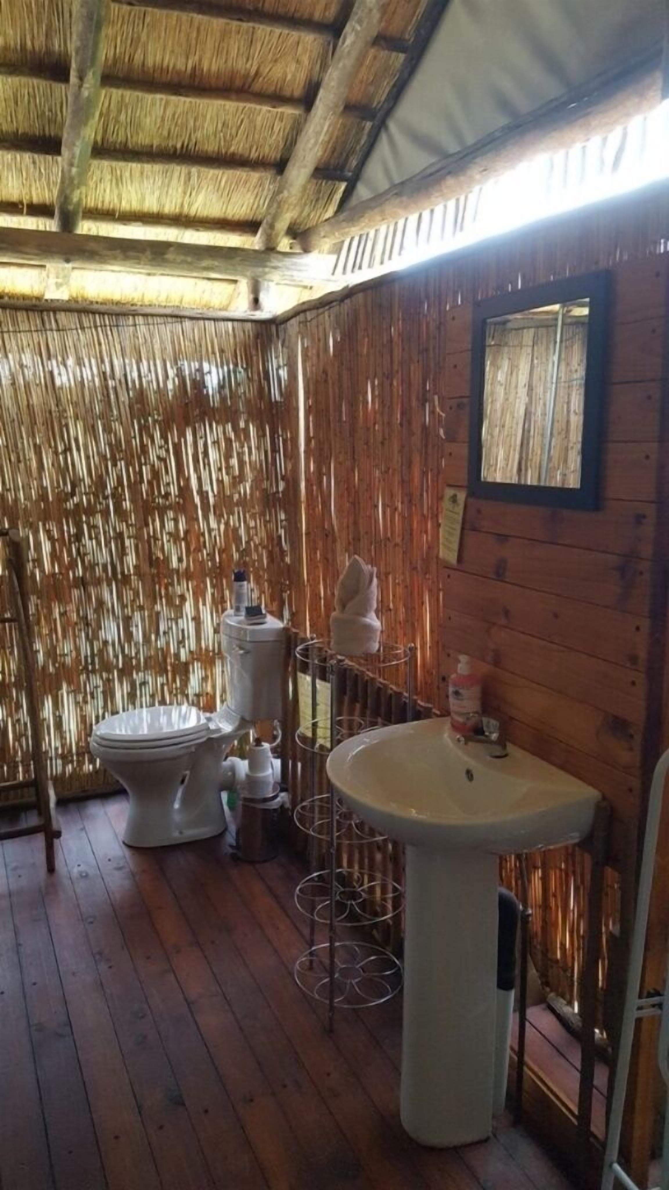 A wooden cabin bathroom with a thatched roof, a toilet, a sink, and a mirror.