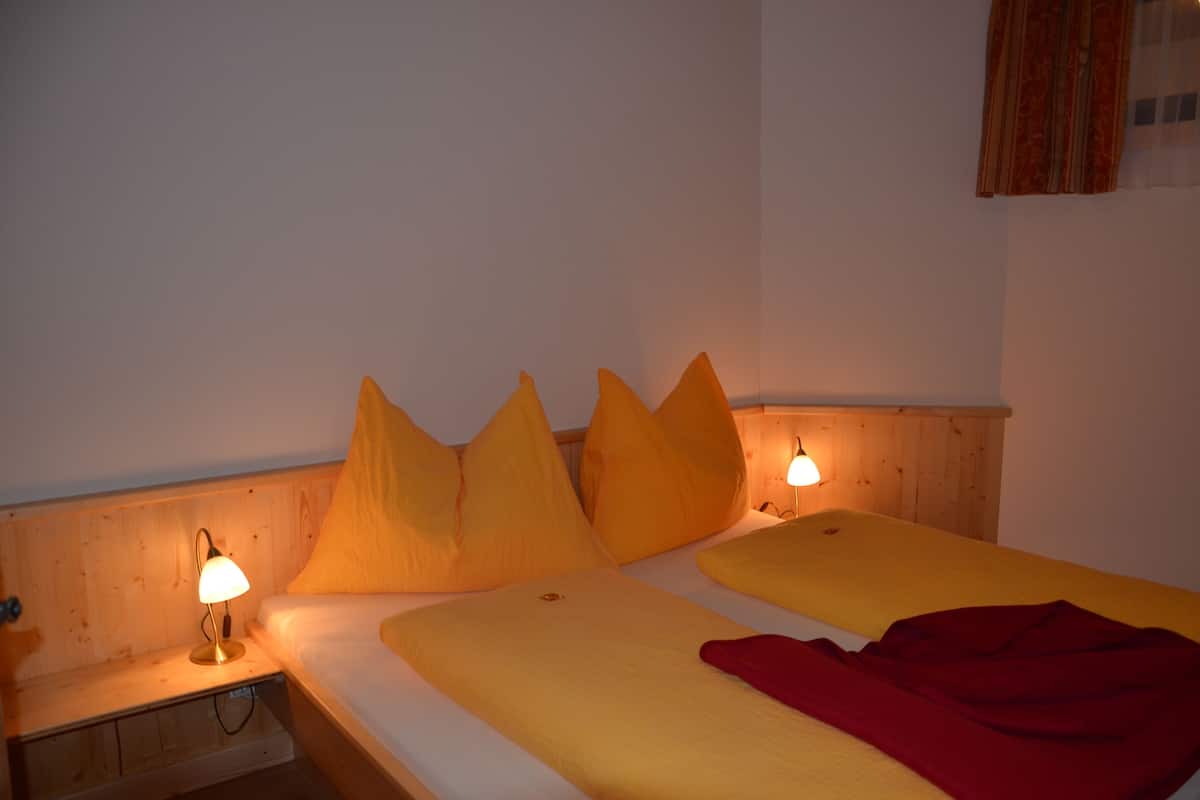 Ein Zimmer mit zwei Betten, jeweils mit gelber Bettdecke und mehreren Kissen. Am linken Bett steht ein Nachttisch mit einer Lampe.