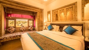 A hotel room with a large bed, a bench with cushions, a view of the desert, and traditional decor. - Hotel Radhika Jaisalmer (Jaisalmer)