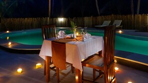 A dining table set for two with candles, surrounded by chairs, in a tropical setting with palm trees and a pool in the background. - Crystal Bay Villa (Penida Island)