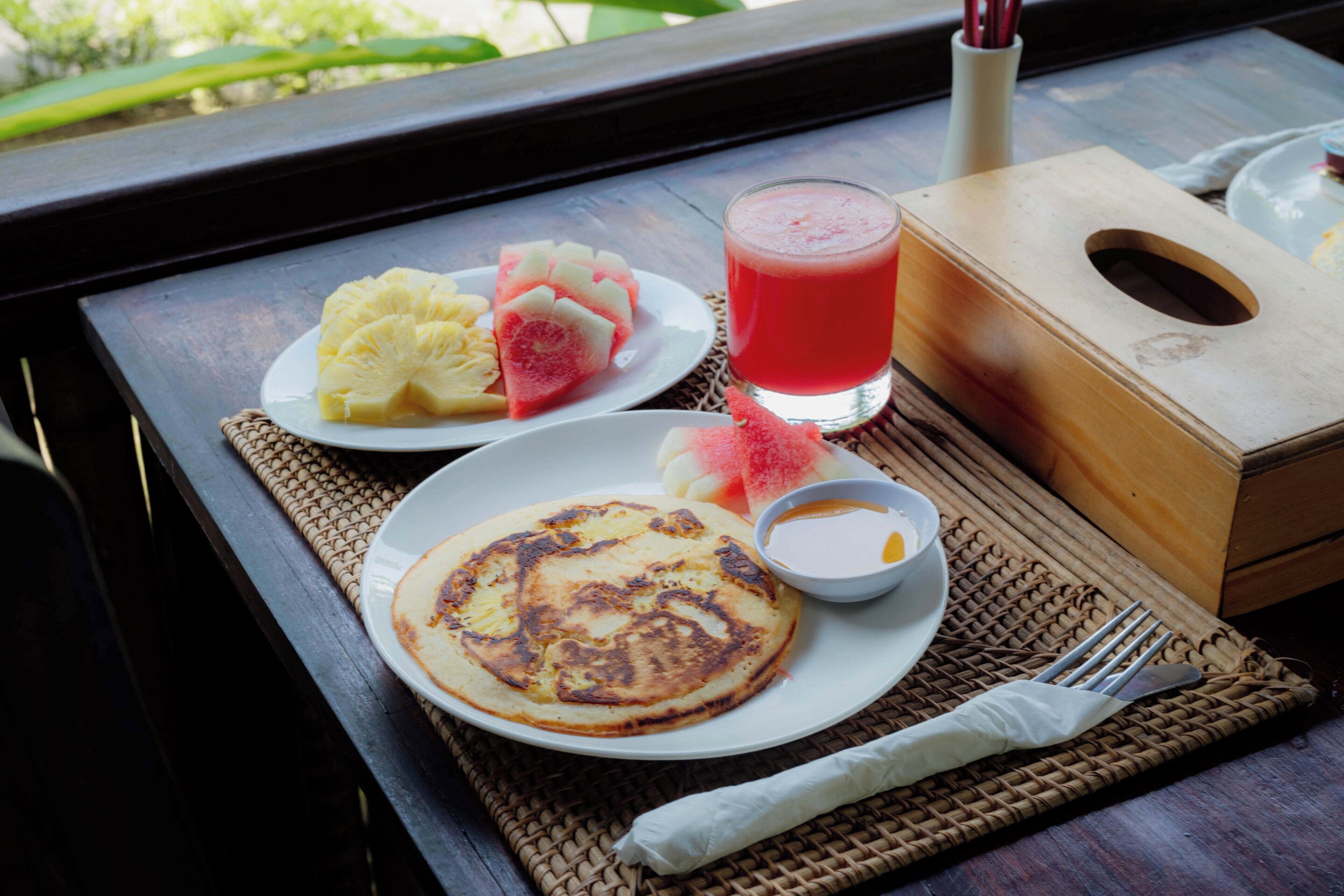 A breakfast tray with pancakes, fruit, and a smoothie on a table with a woven placemat.