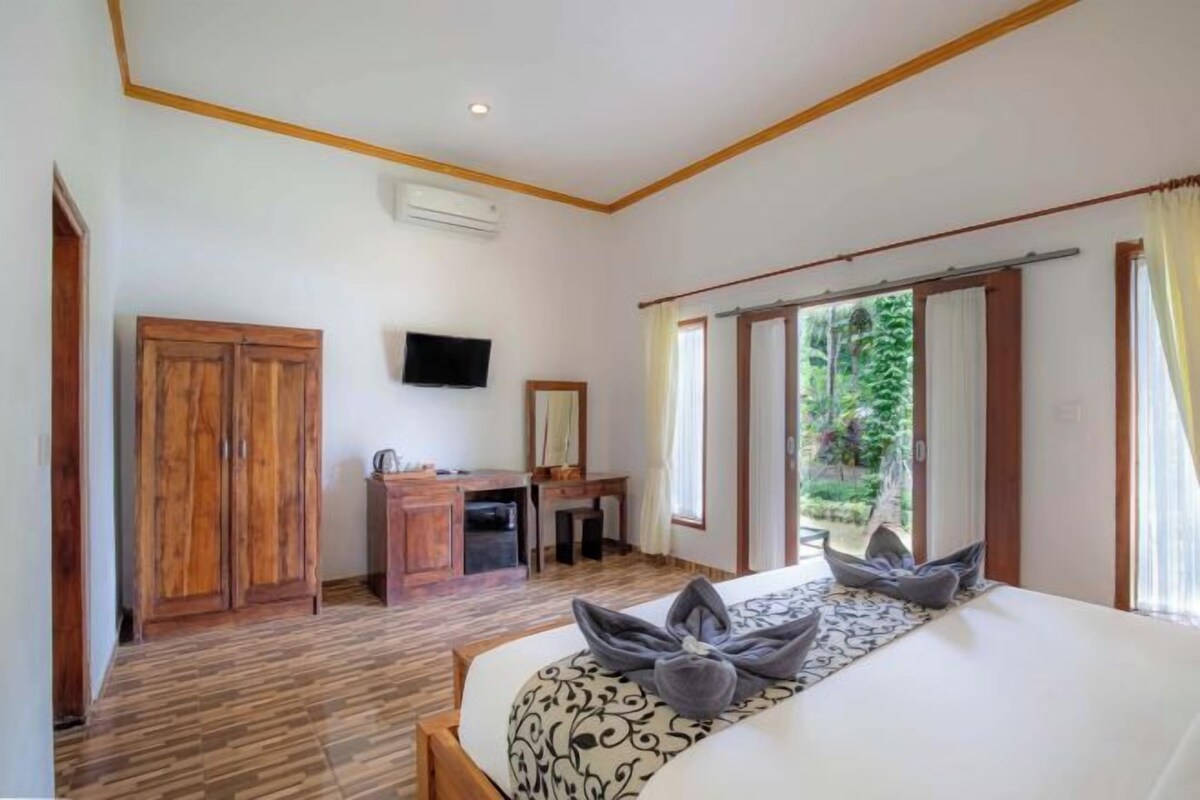A hotel room with a bed, a desk, a television, and a view of greenery outside.