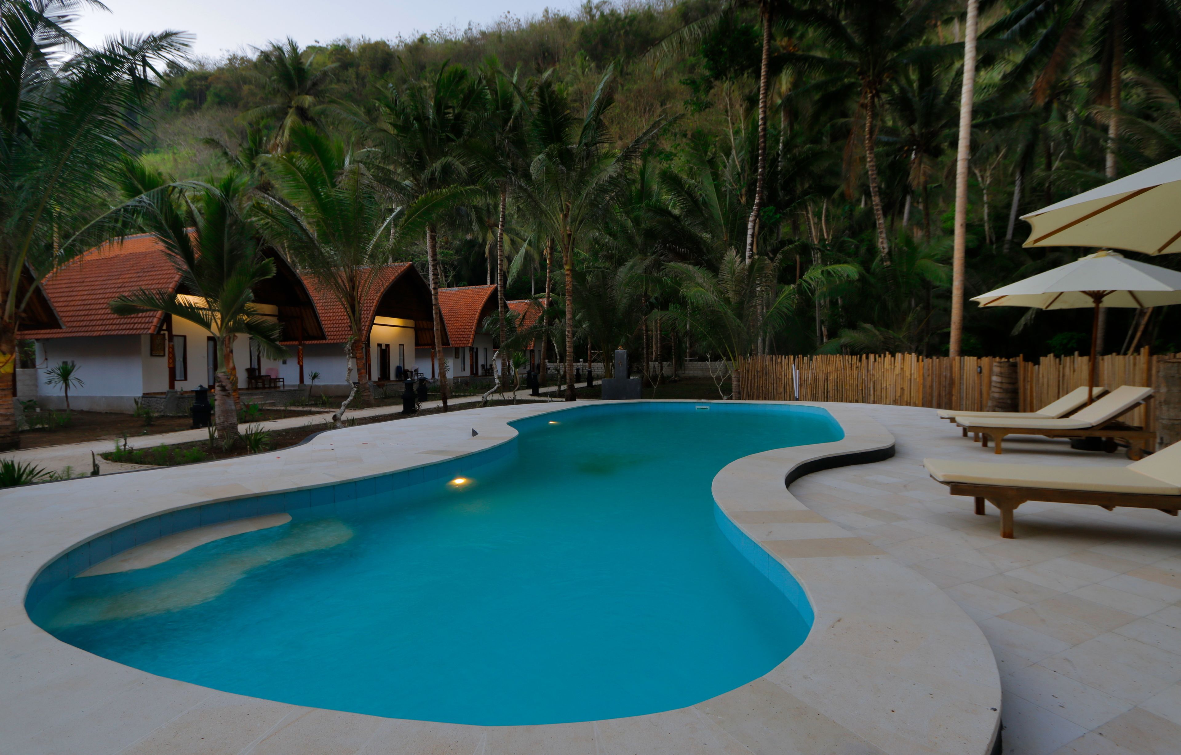 A swimming pool with lounge chairs and umbrellas, surrounded by palm trees and buildings with red roofs.