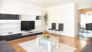 Een moderne woonkamer met een glazen salontafel, grijze banken, een flatscreen televisie en een eethoek met stoelen en een tafel.