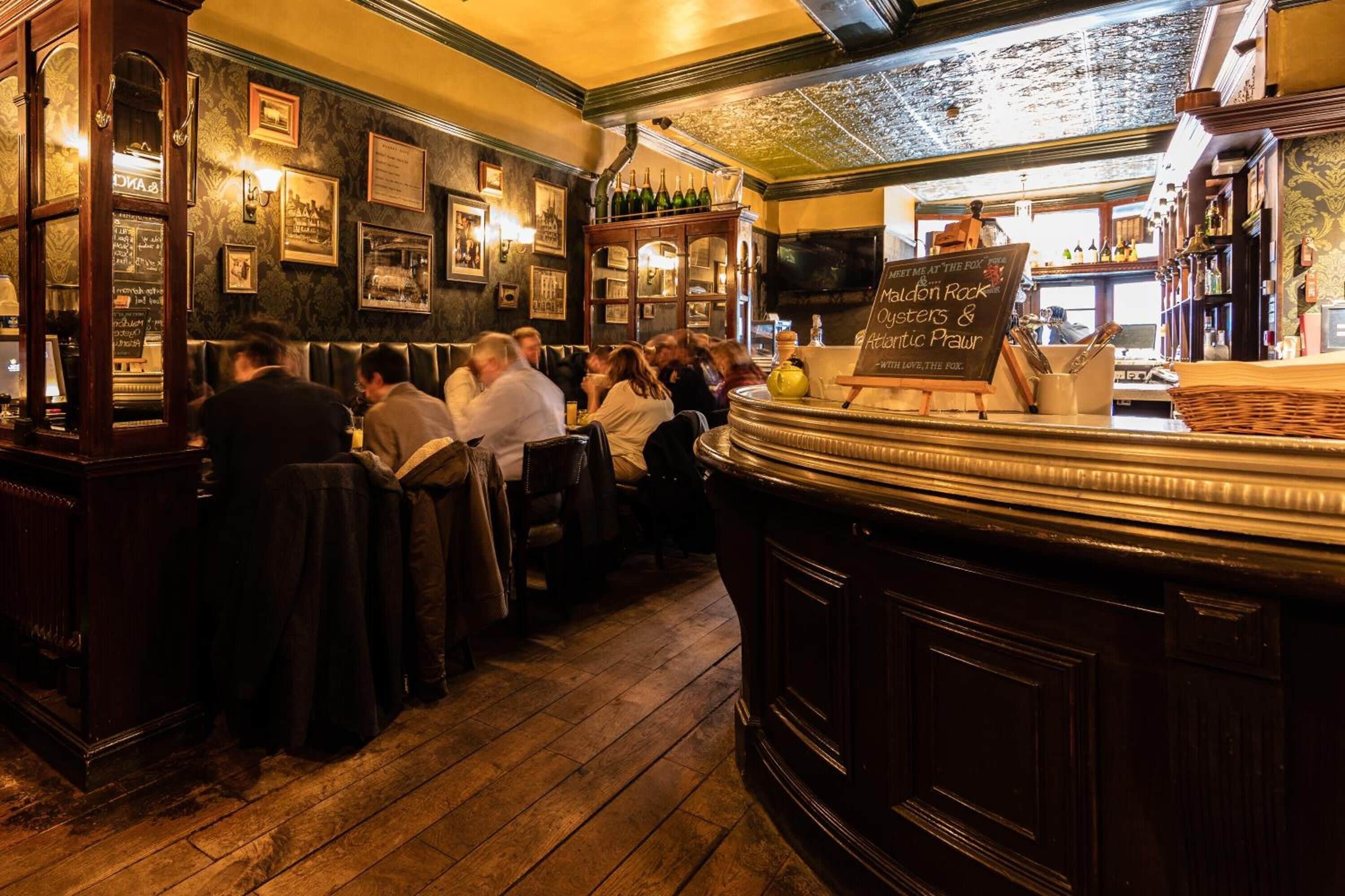 Eine Bar mit Holztischen, einer Kreidetafel mit der Speisekarte und einer Gruppe von Personen, die an den Tischen sitzen.