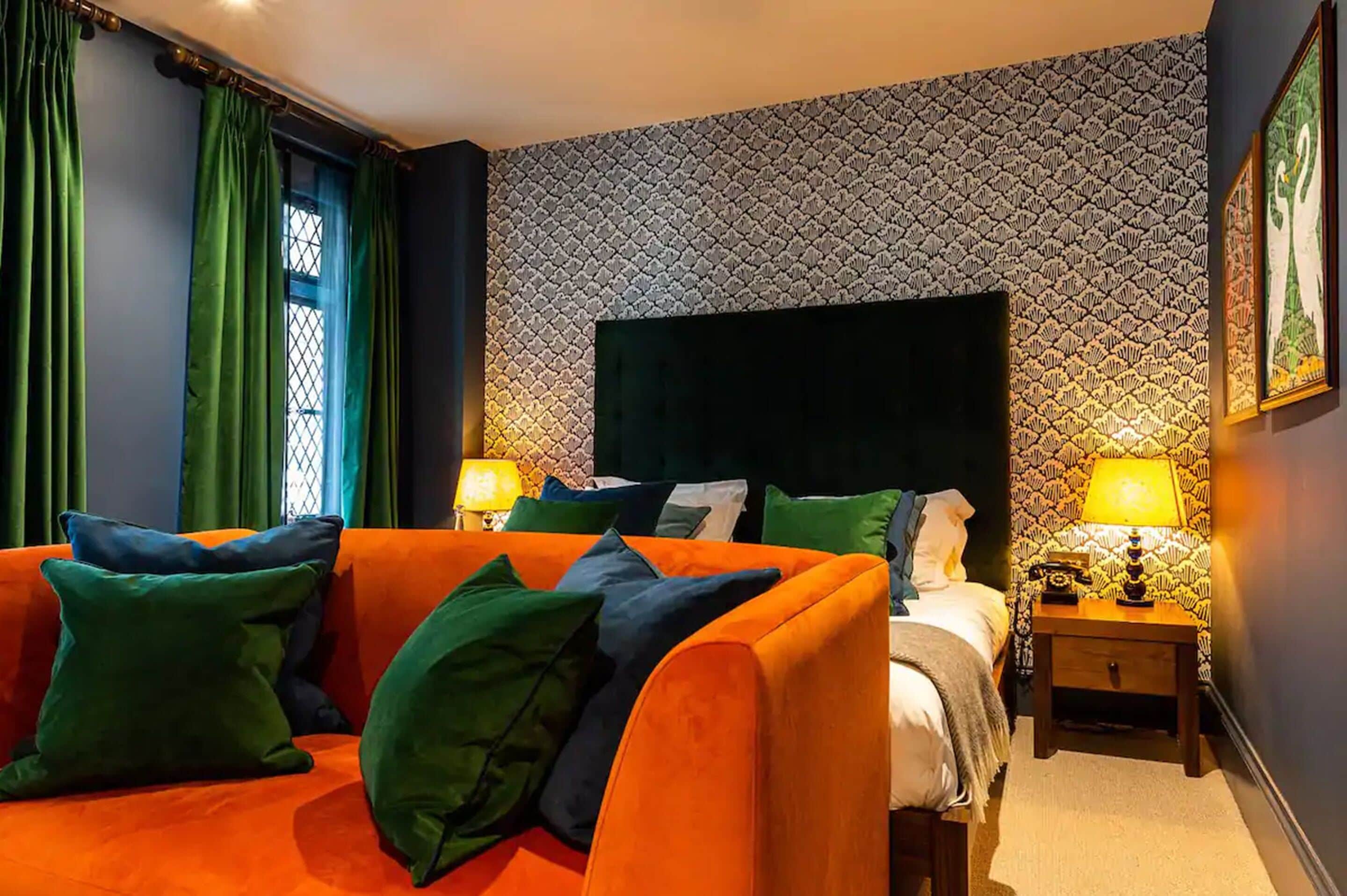 A hotel room with a bed, a sofa, a nightstand with a lamp, and a patterned wall.