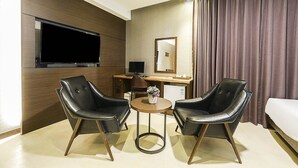 A hotel room with a large flat-screen TV, a desk with a computer, two leather chairs, a small round table with a vase of flowers, and a bed with a white comforter. - Incheon Hotel Bluemarine (Incheon)