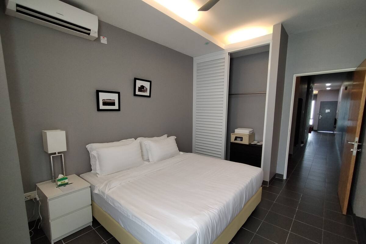 A hotel room with a bed, bedside table, lamp, air conditioning unit, and a door leading to another room.