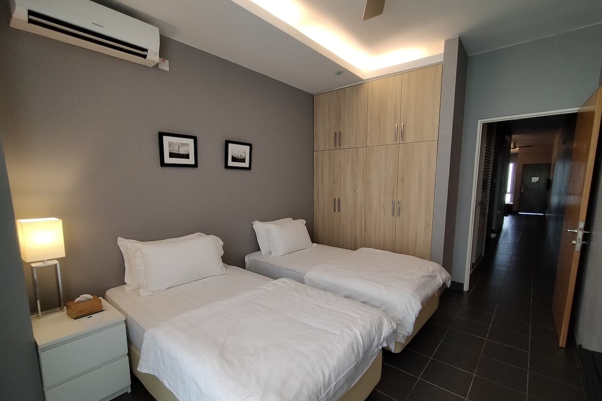 A hotel room with two beds, a bedside table with a lamp, a wardrobe, and a door leading to another room.