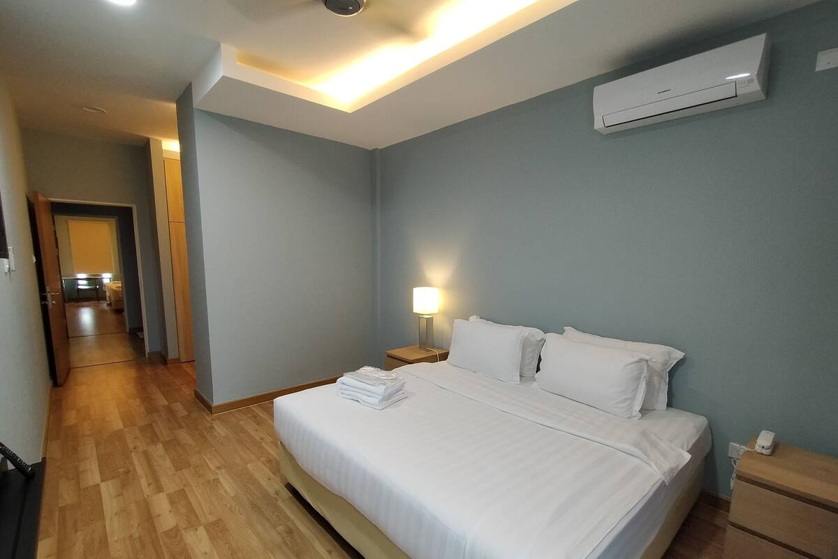 A hotel room with a bed, bedside tables, a lamp, an air conditioner, and a hallway leading to a kitchen area.