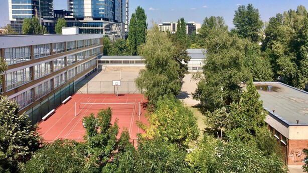 A basketball court surrounded by trees and buildings, including a modern high-rise and a school building.