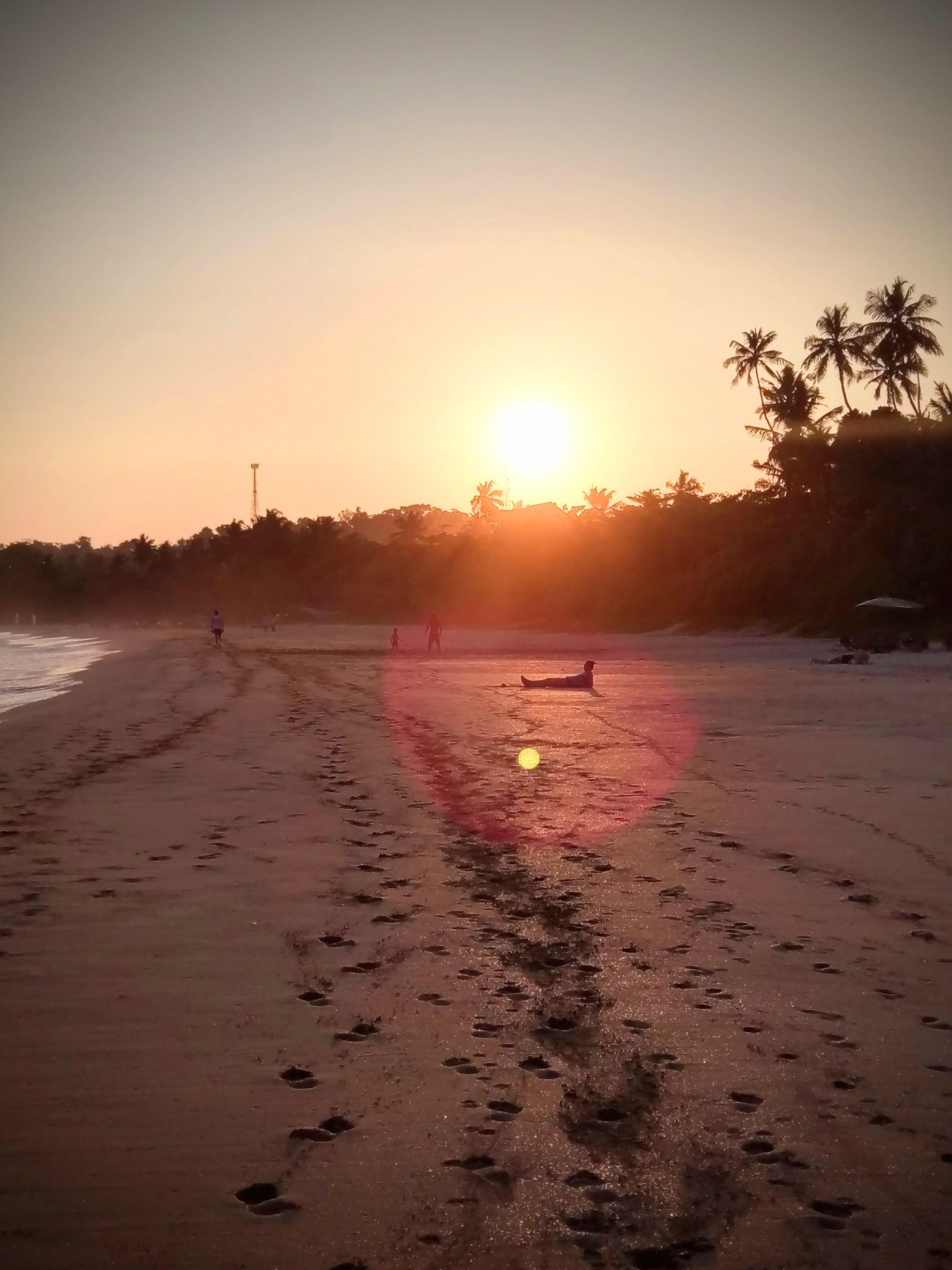 Ein Strand mit Fußspuren, die zum Wasser führen, Palmen im Hintergrund und eine untergehende Sonne am Horizont.