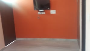 A room with an orange wall, a flat-screen TV mounted on the wall, a bed with white bedding, and a green mat on the floor. - Kukas Guest House (Amer)