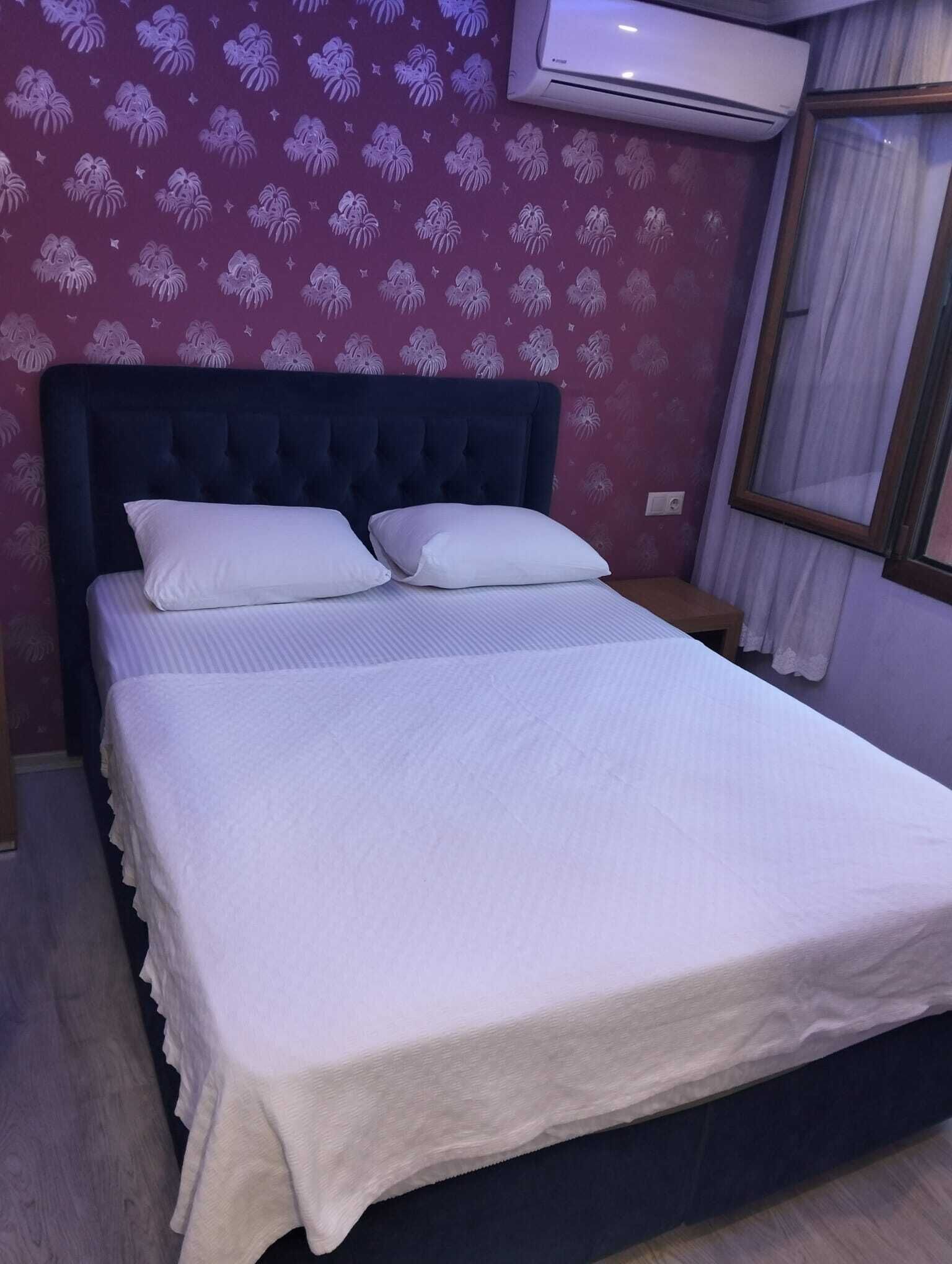 Una cama bien hecha con sábanas blancas, cabecera oscura y pared decorada.