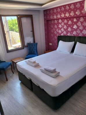 A hotel room with a large bed, a window, a blue chair, and a small table. - Otel Safran Palas (Foça)