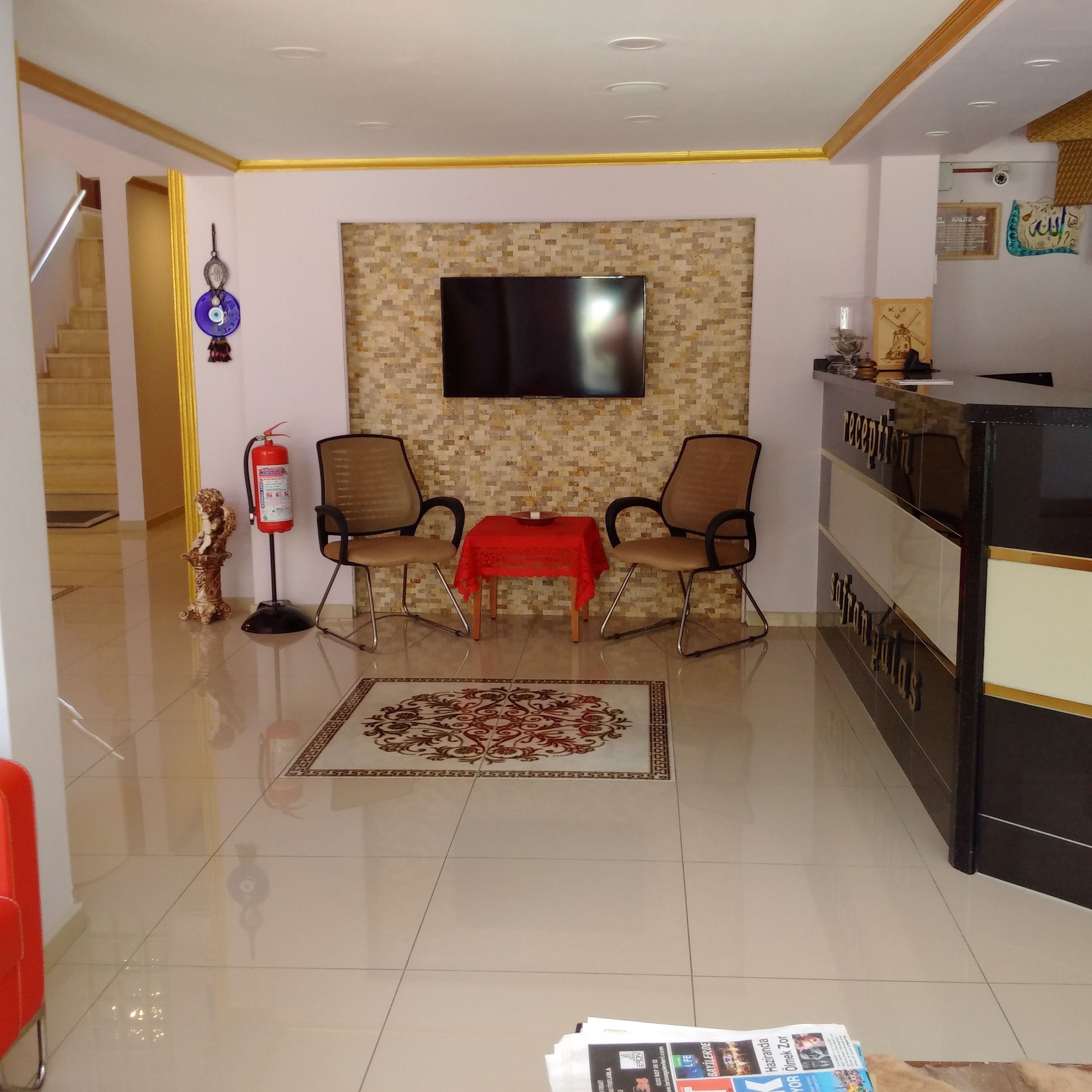 A reception area with a reception desk, two chairs, a red table, a fire extinguisher, a wall-mounted television, and a patterned rug.