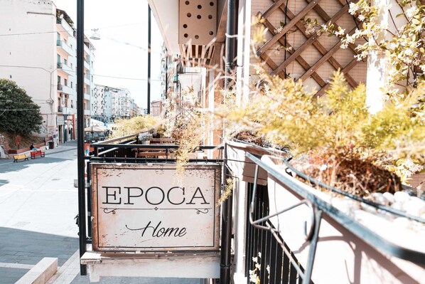 A sign for 'EPOCA Home' with a view of a street lined with buildings and some greenery. - Epoca Home (Cosenza)
