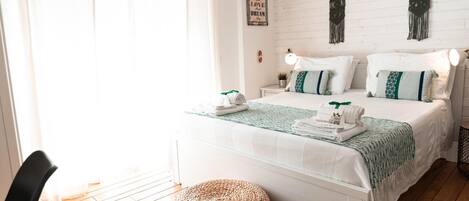 Un dormitorio luminoso y bien iluminado con una cama blanca, ropa de cama verde y blanca, un otoman tejido y una pared decorada con macramé.