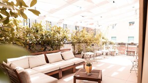Un patio soleado con un sofá de madera y una mesa de centro, rodeado de plantas en macetas y mobiliario de exterior.