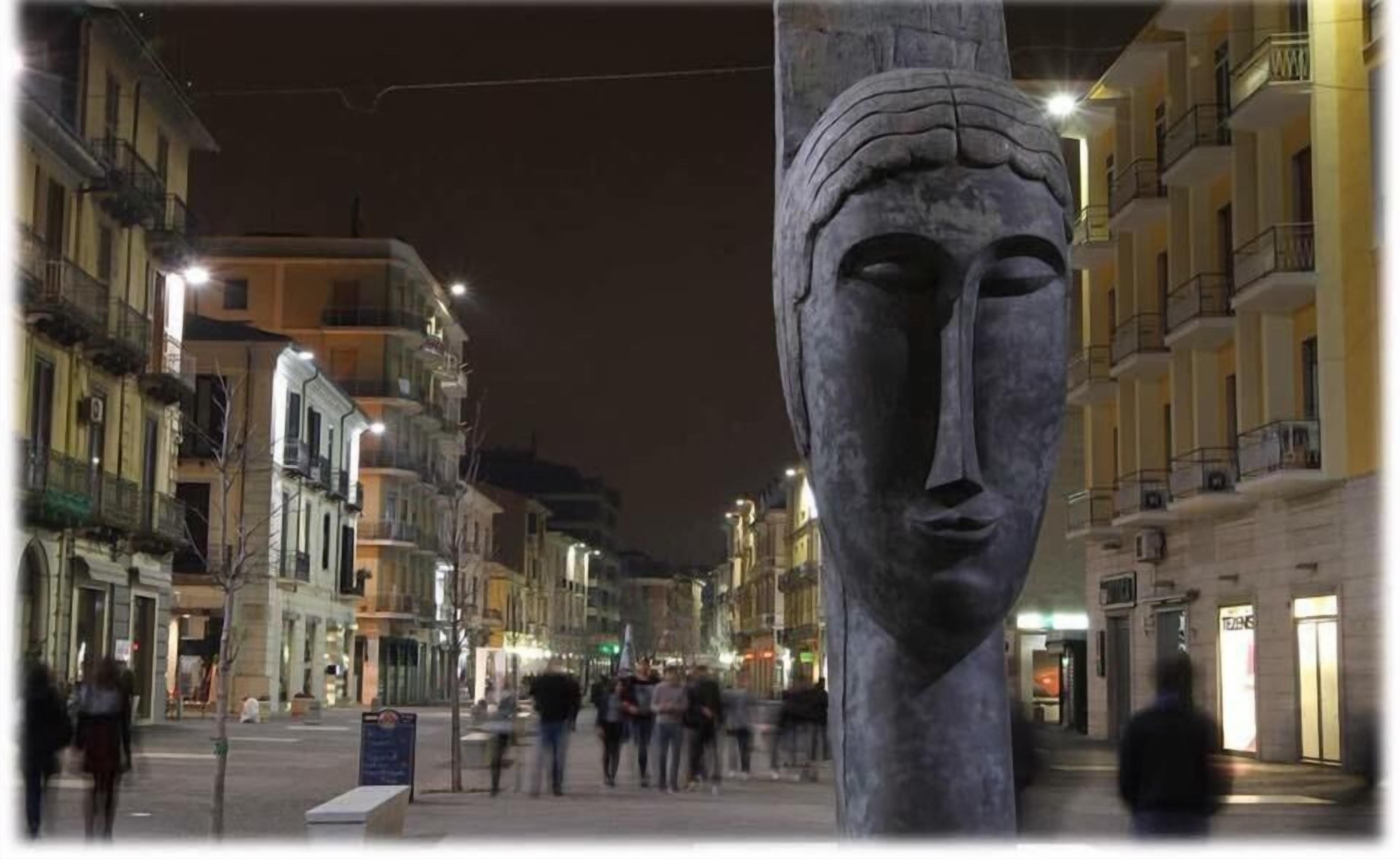 Una escena nocturna en una calle urbana con una destacada escultura de piedra en primer plano.