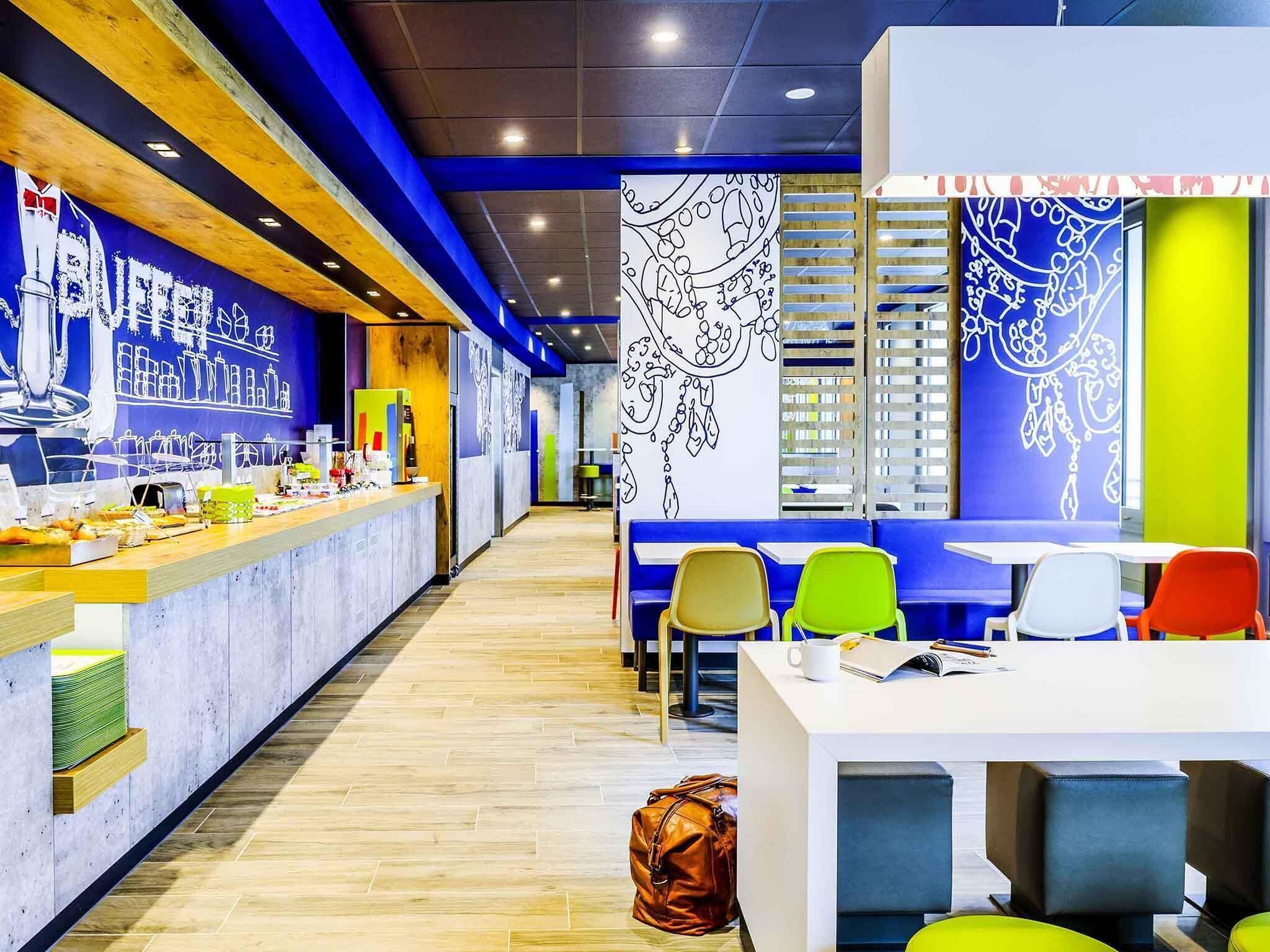Un café moderno con una barra alargada, sillas coloridas y una pared con arte abstracto.
