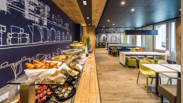 A modern cafe with a long counter displaying various food items, wooden ceiling, and colorful seating. - ibis budget Frankfurt City Ost (Frankfurt)