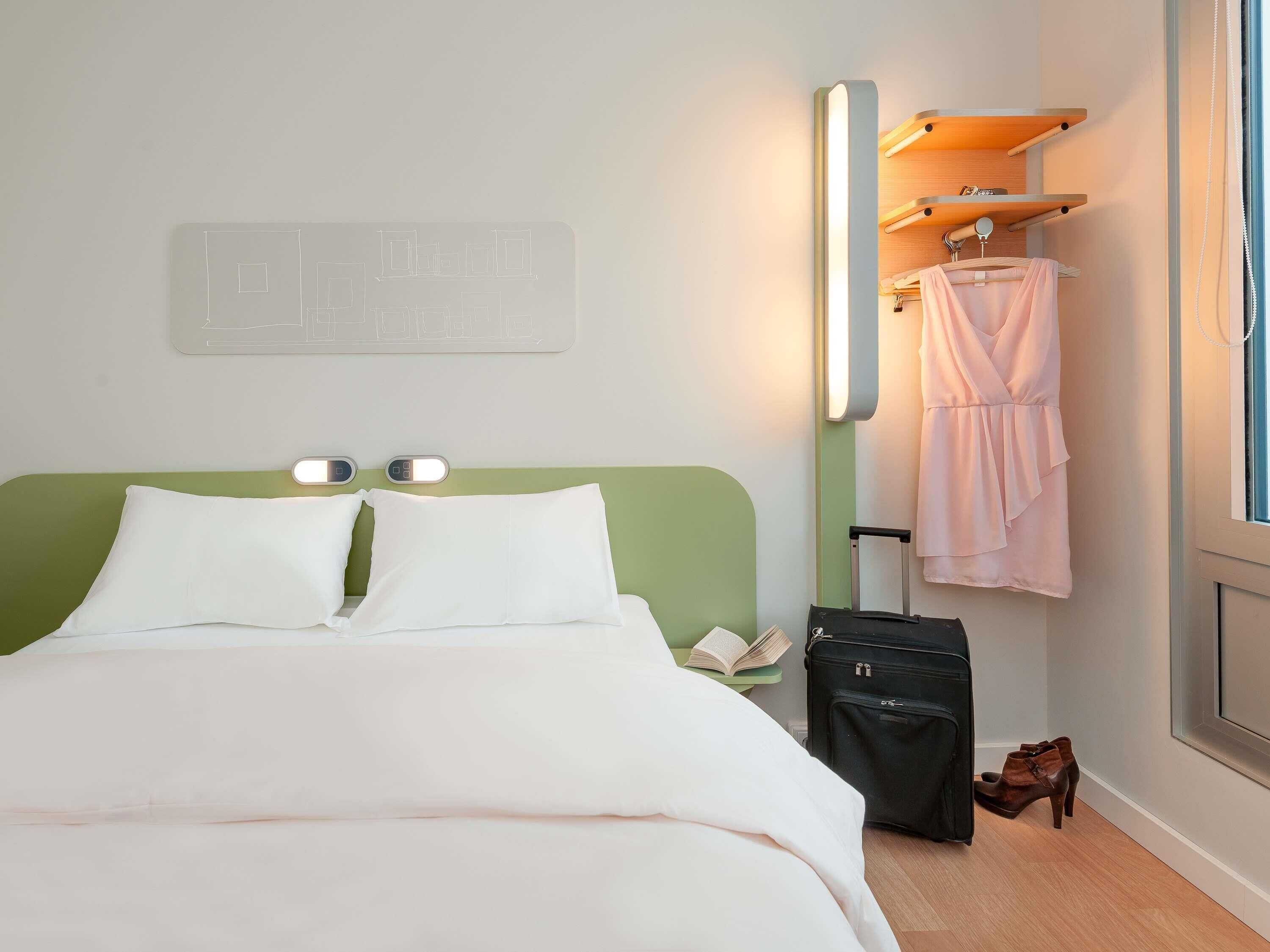 Una camera d'albergo con una testiera verde, un letto con biancheria bianca, un comodino con una lampada, una valigia e un accappatoio appeso a un appendiabiti di legno.