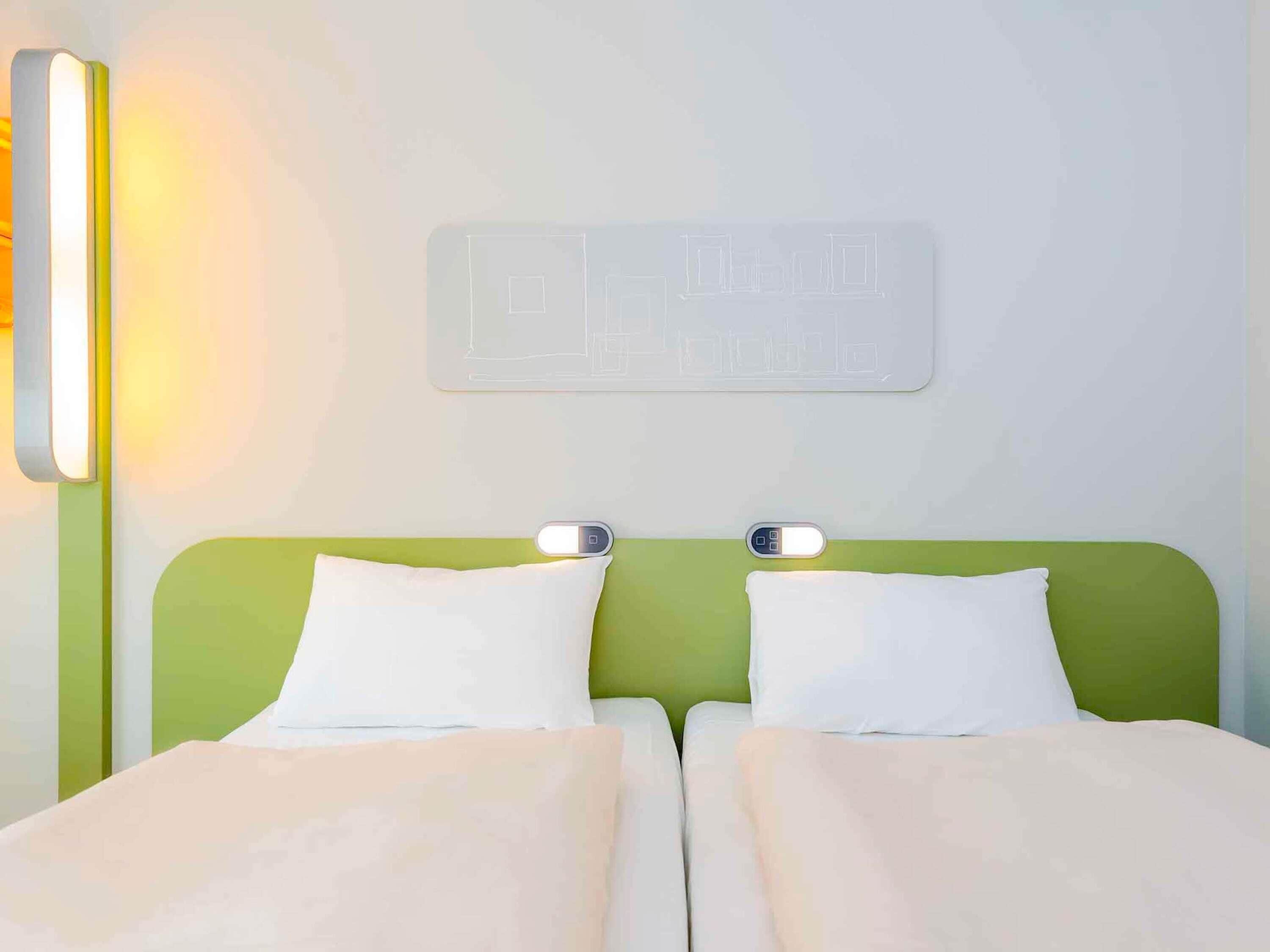 Une chambre d’hôtel avec une tête de lit verte, deux oreillers blancs et un luminaire fixé au mur.